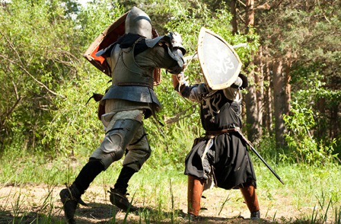 Kaksi, keskiaikaiseen kevyeen haarniskaan pukeutunutta henkilöä taistelemassa metsässä miekoin ja kilvin.